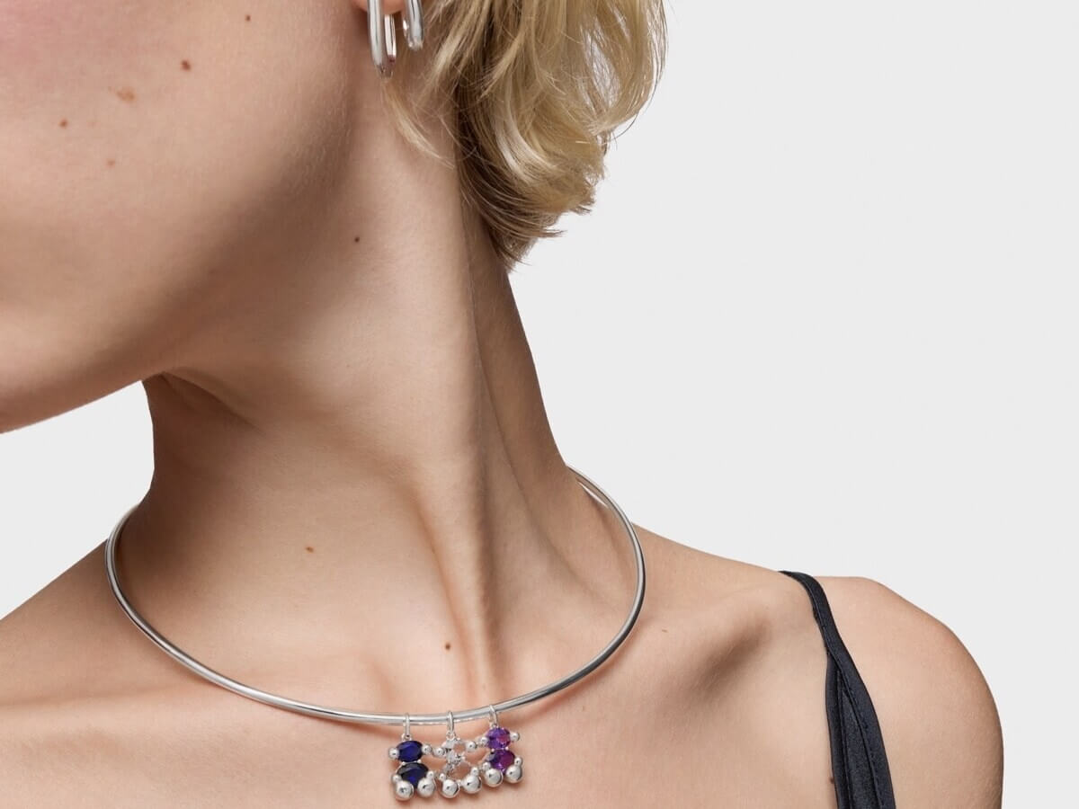 Kobieta ze sztywnym, srebrnym naszyjnikiem na szyi. Naszyjnik ma zawieszki z szafirem w kształcie misia TOUS