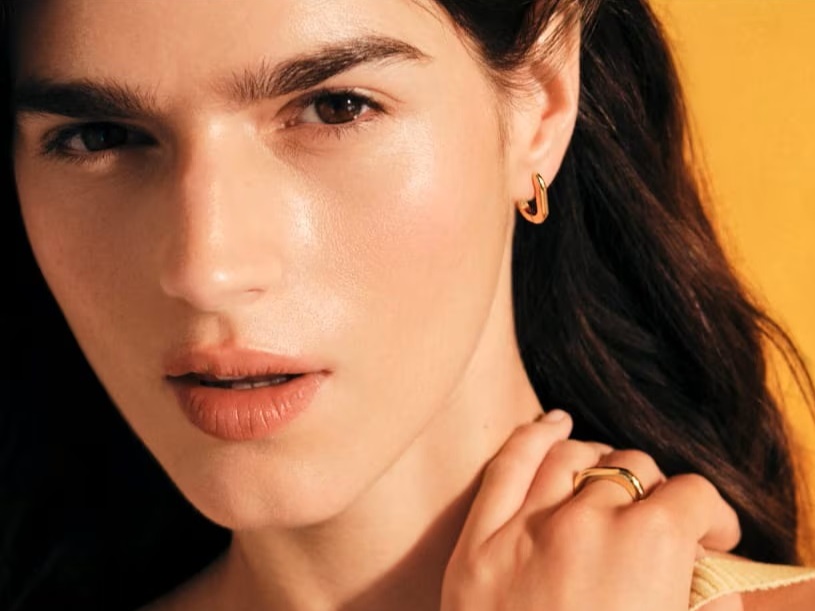 Twarz ciemnowłosej kobiety na złotym tle, kobieta ma na uszach złote kolczyki TOUS